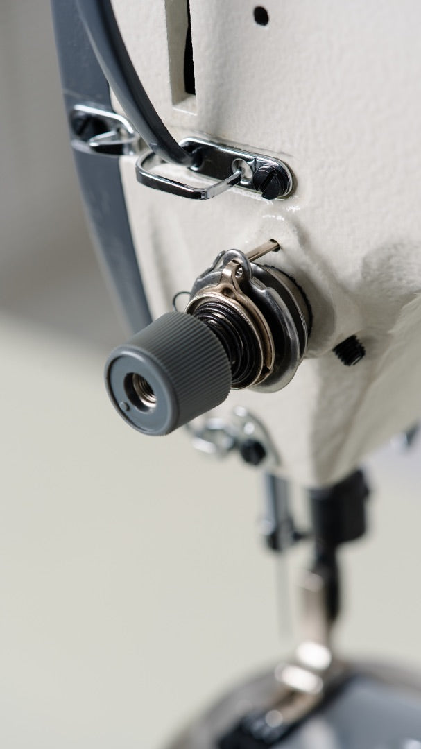 Close-up of industrial wig sewing machine tension dial, thread spring, and thread guides on the upper thread path